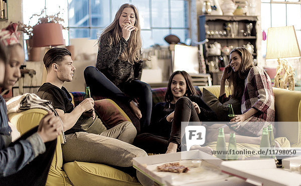 Vier junge Frauen und ein junger Mann sitzen lächelnd auf einem Sofa  Pizza und Bierflaschen auf dem Couchtisch.
