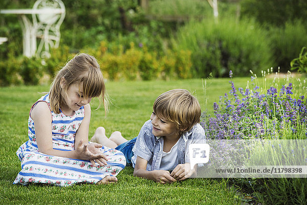 A girl sitting on the grass talking to her brother lying beside her on a lawn in a garden.
