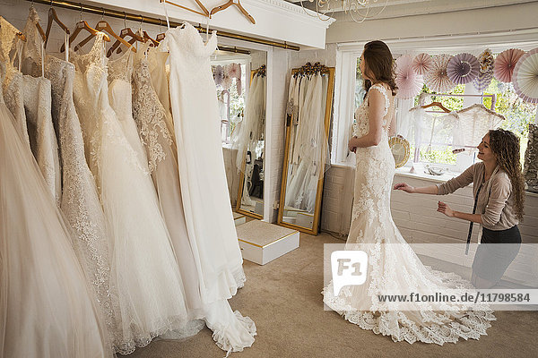 Ausgestellte Reihen von Brautkleidern. Eine junge Frau in einem weißen Hochzeitskleid in voller Länge  die ihr Spiegelbild im Spiegel in einer Brautboutique betrachtet.