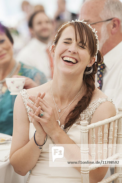 Braut im Hochzeitskleid im Festzelt sitzend  die Hände gefaltet und lachend.