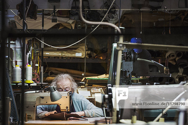 Eine ältere grauhaarige Arbeiterin sitzt an einer Ledernähmaschine in einer Schusterwerkstatt.