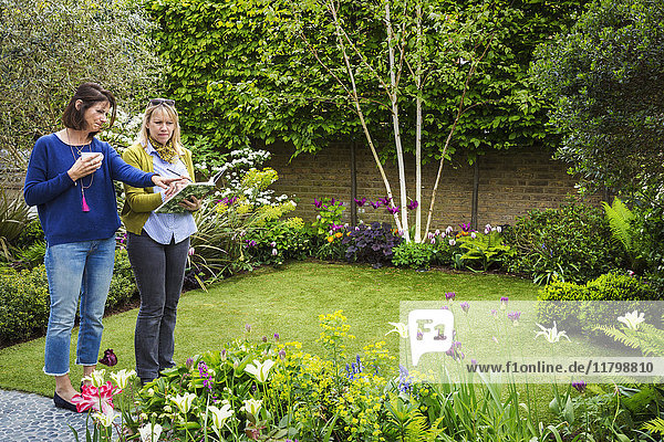 Zwei Frauen stehen in einem Garten auf einer von Blumenbeeten umgebenen Wiese und diskutieren über die Gartengestaltung.