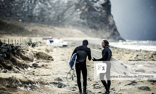 Two surfers wearing wetsuits and carrying surfboards walking towards a van.