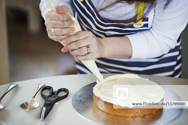 Close up of person wearing a blue and white stripy apron  holding a piping bag  decorating a cake with cream  spoons and scissors on table.