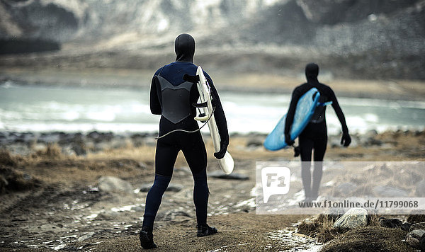 Two surfers wearing wetsuits and carrying surfboards walking towards the ocean with mountains behind.