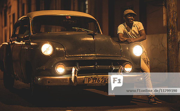 Mann  der sich nachts in einer Straße an einen Oldtimer aus den 1950er Jahren lehnt.