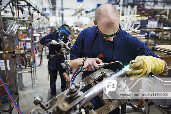 Männlicher Fabrikfacharbeiter mit Schutzbrille  der ein Schweißwerkzeug benutzt und in einer Fabrik auf einem Fahrrad arbeitet.