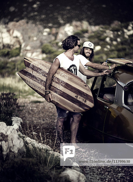 Zwei junge Männer stehen neben dem Auto und tragen ein Surfbrett.