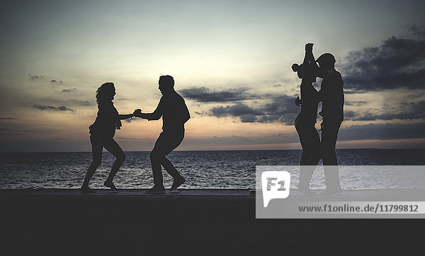 Four people dancing on a sea wall in front of the ocean at dusk.