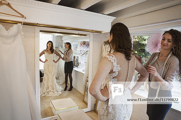 Eine junge Frau in einem weißen Hochzeitskleid in voller Länge  die ihr Spiegelbild im Spiegel in einer Brautboutique betrachtet. Eine Schneiderin im Hintergrund.