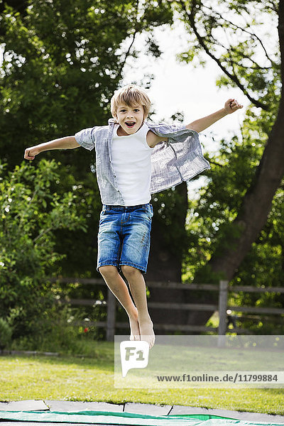Junge in Hemd und Jeans-Shorts  der auf einem Trampolin in einem Garten springt.