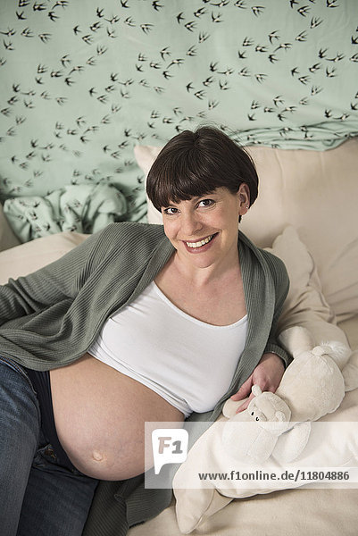 Porträt einer lächelnden schwangeren Frau mit Plüschtier auf dem Bett liegend