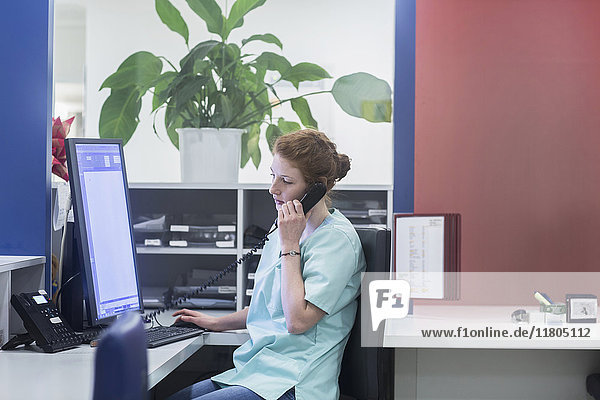 Krankenschwester bei der Arbeit am Schwesternarbeitsplatz  während sie einen Computer benutzt und telefoniert