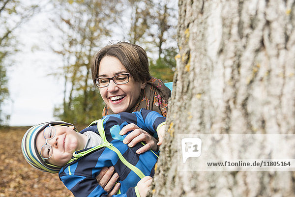 Mutter und Sohn spähen von einem großen Baum und lachen in der Herbstlandschaft