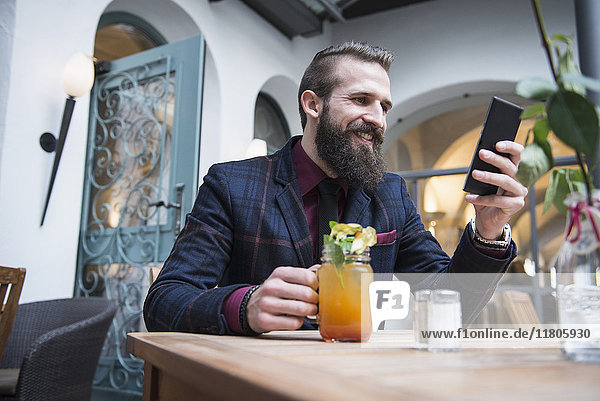 Junger Mann  der ein Mobiltelefon benutzt und ein Mocktail-Glas hält