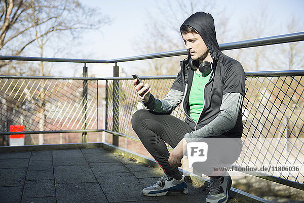 Mann in Sportkleidung mit Kopfhörern und Smartphone entspannt sich nach dem Training auf einer Brücke