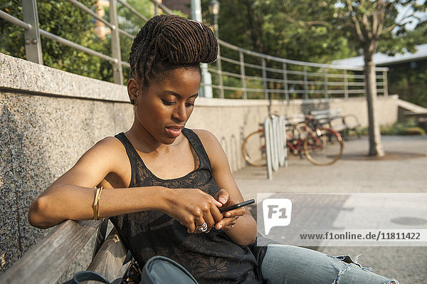 Afroamerikanische Frau auf einer Bank im Park  die eine SMS auf ihrem Handy schreibt