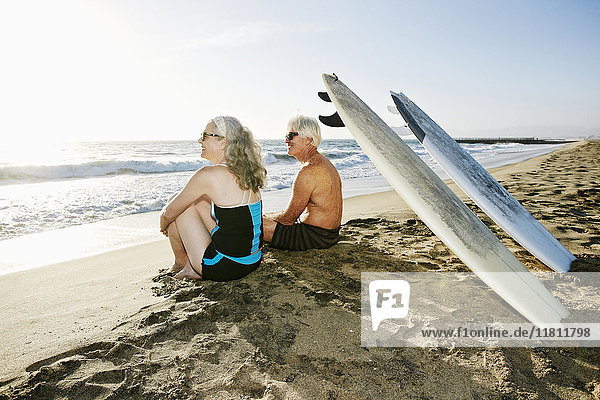 Older Caucasian couple sitting on beach with surfboards