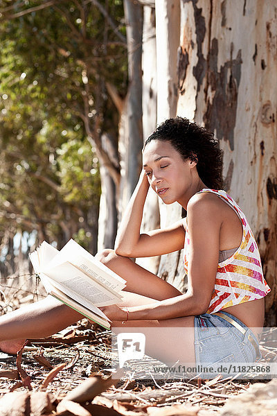 Junge Frau sitzt am Fuße eines Baumes und liest ein Buch