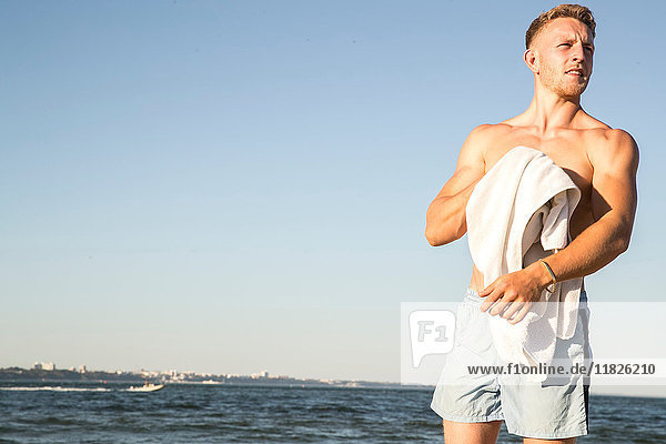 Junger männlicher Schwimmer trocknet sich mit Handtuch am Strand
