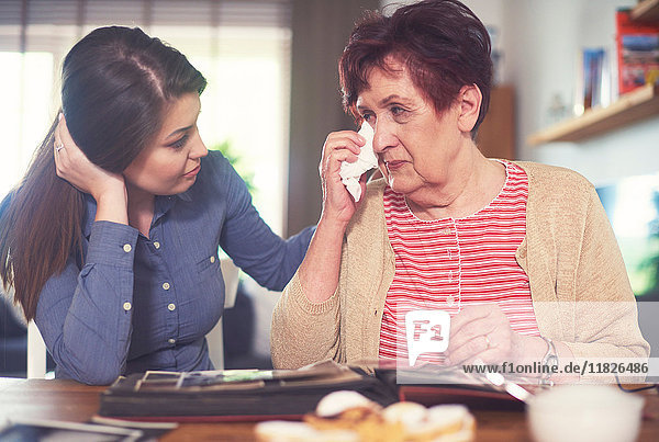 Junge Frau bei Tisch mit Großmutter weint beim Betrachten des Fotoalbums