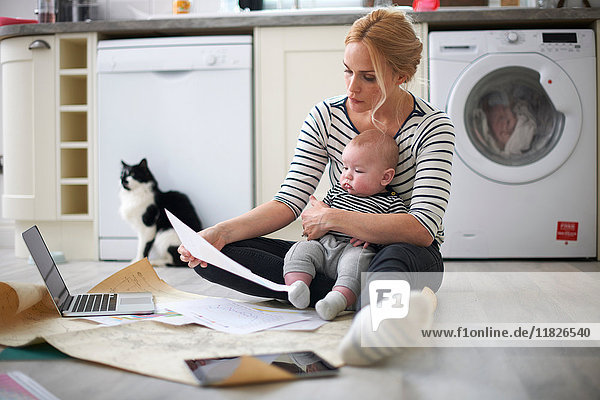 Frau hält kleinen Sohn im Arm und schaut durch die Arbeit auf dem Küchenboden  Laptop und digitales Tablett vor sich
