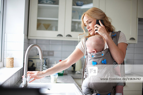 Frau hält kleinen Jungen im Tragetuch  benutzt Smartphone  dreht Wasserhahn auf  um den Abwasch zu erledigen