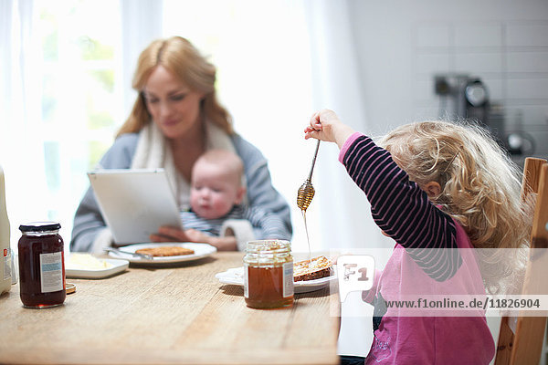 Mutter hält einen kleinen Jungen in der Hand  schaut auf ein digitales Tablett  sitzt mit der kleinen Tochter am Küchentisch  frühstückt