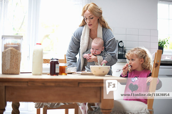 Mutter  die einen kleinen Jungen hält  mit der kleinen Tochter am Küchentisch sitzt und frühstückt