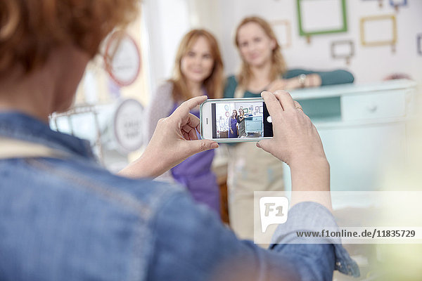 Frau mit Fotohandy beim Fotografieren von Klassenkameraden, die am gemalten Beistelltisch in der Kunstklasse posieren.