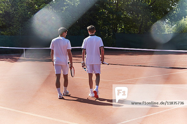 Junge Tennisspieler, die mit Tennisschlägern auf einem sonnigen Sandplatz spazieren gehen.
