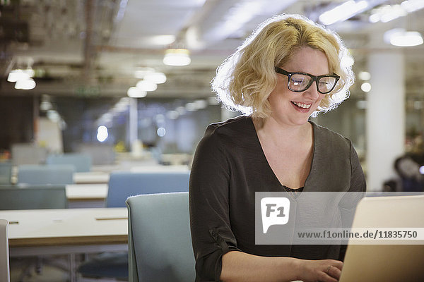 Lächelnde Geschäftsfrau arbeitet spät am Laptop im dunklen Büro