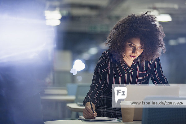 Fokussierte Geschäftsfrau  die spät am Laptop arbeitet und Notizen im dunklen Büro macht.