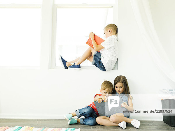Boy (6-7) reading book and sister with brother (2-3  6-7) using digital tablet