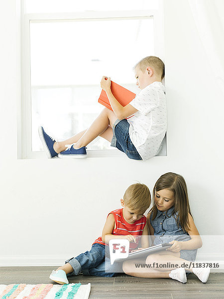 Boy (6-7) reading book and sister with brother (2-3  6-7) using digital tablet