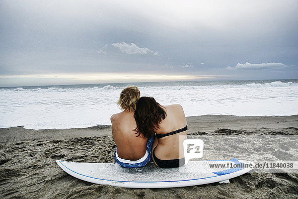 Kaukasisches Paar sitzt auf einem Surfbrett am Strand