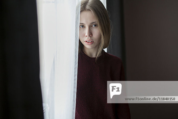 Serious Caucasian woman standing near curtain at window