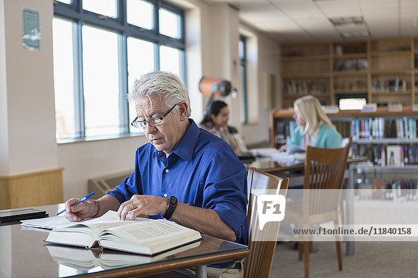 Älterer Mann liest ein Buch in der Bibliothek und schreibt Notizen