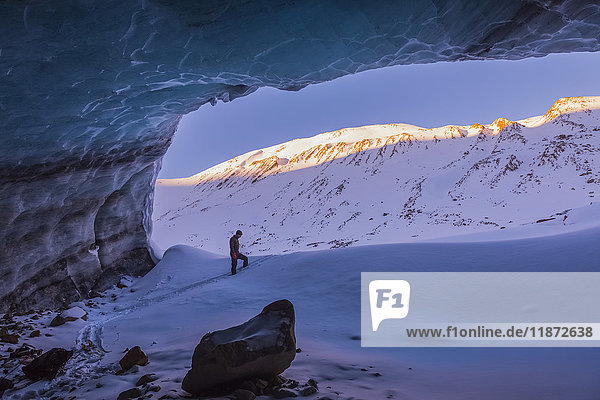 Ein Mann und die Gipfel der umliegenden Berge bei Sonnenaufgang werden vom Eingang zu einer großen Eishöhle im Augustana-Gletscher in der Alaska Range im Winter eingerahmt  Inner-Alaska  USA