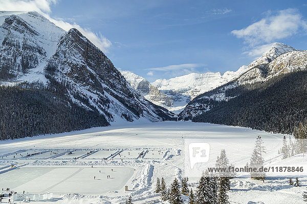 'A wide angle view of Lake Louise from the upper floor of Chateau Lake Louise Resort in the winter surrounded by snow capped mountains and filled with skaters on the ice rinks carved into the frozen lake; Lake Louise  Alberta  Canada'