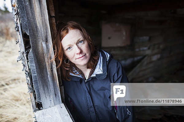 'Portrait of a woman with red hair leaning against a wooden wall and looking up at the camera; Alaska  United States of America'