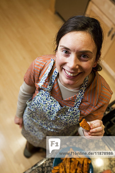 Hochformatige Ansicht einer Frau in einer Küche  die eine Schürze trägt und ein Stück gebackene Süßkartoffel hält; Alaska  Vereinigte Staaten von Amerika'.
