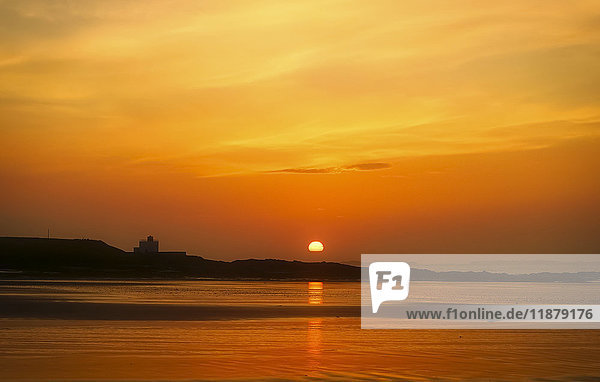 'The golden sun sinking on the horizon with a silhouette of the coastline and an orange sky reflected in the water; Bamburgh  Northumberland  England'
