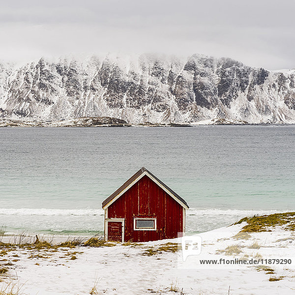 Eine einsame rote Hütte am Wasser mit schroffen  schneebedeckten Bergen auf der anderen Seite des Wassers; Lofoten  Nordland  Norwegen'.