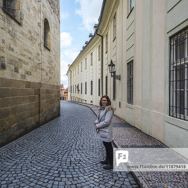 'A woman stands posing on a narrow street with patterned paving stones in between buildings; Prague  Czech Republic'
