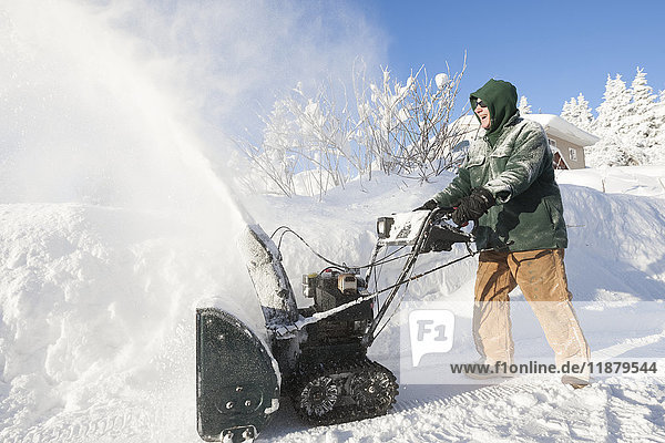 Ein Mann benutzt fröhlich seine Schneefräse vor seinem Haus; Alaska  Vereinigte Staaten von Amerika'.