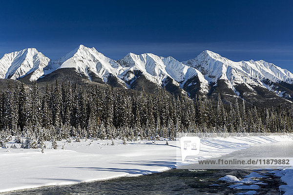 Schneebedeckte Bergkette und immergrüne Bäume entlang eines schneebedeckten Flussufers und ein offener Fluss mit blauem Himmel; Radium Hot Springs  British Columbia  Kanada'.