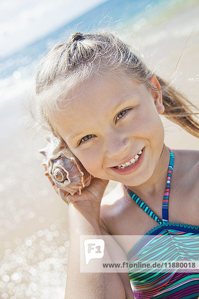 Ein junges Mädchen lächelt  hält eine Muschel in der Hand und lauscht dem Klang einer Muschel an ihrem Ohr am Strand; Honolulu  Oahu  Hawaii  Vereinigte Staaten von Amerika'.