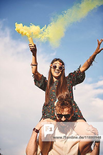 Young boho woman holding yellow smoke flare on boyfriend's shoulders at festival