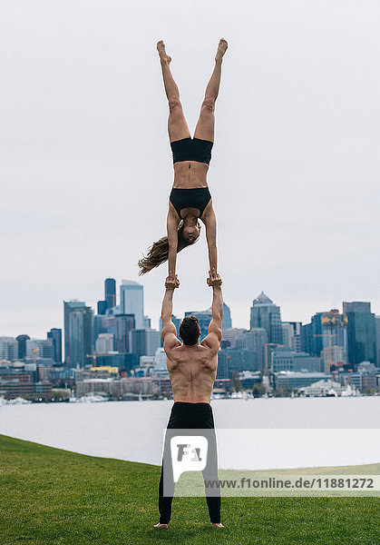 Junge Frau balanciert auf Männerhänden  in voller Länge  praktiziert Yoga vor der Skyline von Seattle